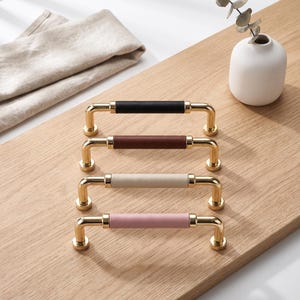 Puede incluir: Cuatro tiradores de armario con herrajes de metal dorado pulido y agarres centrales cilíndricos. Los agarres tienen un acabado en colores mate sólidos: negro, marrón oscuro, beige y rosa empolvado. Cada tirador tiene dos bases de montaje.
