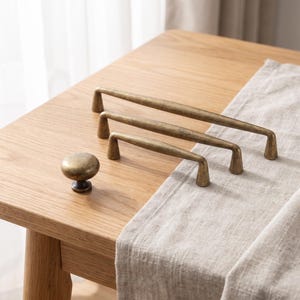 May include: A set of three antique brass cabinet pulls of varying lengths and one matching round cabinet knob. Each piece features a smooth, brushed metallic finish with a warm, golden-brown tone and tapered feet for mounting to furniture or doors.