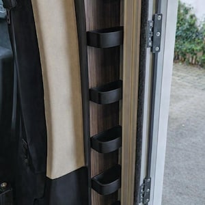 May include: A vertical storage unit featuring a dark wood grain panel with five matte black, semi-circular shelves mounted in a column. The unit is positioned next to a beige upholstered panel and a black fabric section, with a metal hinge visible.
