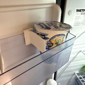 May include: A container of cottage cheese sits inside a refrigerator shelf. The container is white with blue and yellow text. The refrigerator is white and has clear plastic shelves.