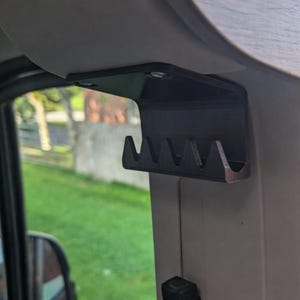 May include: Black metal accessory holder with multiple slots, mounted inside a vehicle. The holder is attached to the interior trim near the window and appears to be designed for organizing small items. The background shows a view of the outdoors.