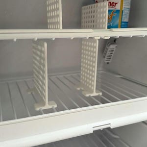 Op de afbeelding: Witte plastic koelkastplankverdelers met een geperforeerd ontwerp. De T-vormige verdelers zijn ontworpen om items op de koelkastplanken te organiseren. Een bakje boter is zichtbaar op de bovenste plank.