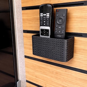 Pode incluir: Um suporte preto para controle remoto em estilo tecido com dois controles remotos pretos. O suporte é montado em um painel de madeira marrom claro com detalhes horizontais pretos. Os controles remotos têm vários botões e controles.