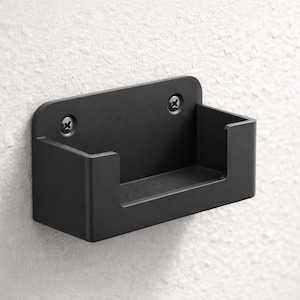May include: A black metal wall-mounted holder with a rectangular design. The holder is attached to a white wall with two screws. The holder has a U-shaped opening.