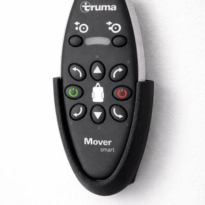 Könnte beinhalten: Eine schwarz-weiße Truma Mover Smart-Fernbedienung mit mehreren Tasten und einem Halter. Die Fernbedienung hat eine Vielzahl von Symbolen und das Wort "Truma" oben. Die Fernbedienung ist an einer weißen Wand befestigt.