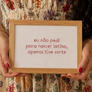 May include: A rectangular wooden frame containing a white fabric panel with red embroidered text that reads: eu não pedi para nascer latina, apenas tive sorte. The frame is held against a cream-coloured dress featuring a vibrant floral pattern.