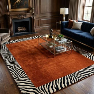 May include: A rectangular area rug featuring a solid burnt orange centre panel surrounded by a wide border with a black and white zebra stripe pattern. The rug is positioned on a dark wood floor in a room with a fireplace and blue sofa.