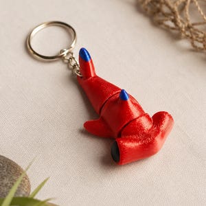 May include: A red hammerhead shark keychain with a metallic silver ring. The shark figure features a textured surface, blue accents on the dorsal and pectoral fins, and black eyes. The design includes a segmented body structure with visible joints.