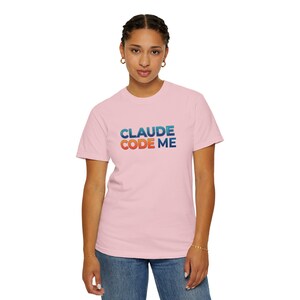 Op de afbeelding: Een lichtroze T-shirt met korte mouwen en de tekst CLAUDE CODE ME op de borst gedrukt. Het woord CLAUDE is in een blauw verlooplettertype, terwijl de woorden CODE ME in een oranje verloop met een donkere omlijning zijn weergegeven.