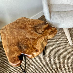 May include: A unique, live-edge wooden side table with a natural, organic shape. The table is made of light brown wood with dark grain patterns and sits on three black metal hairpin legs. The table is approximately 24 inches wide.