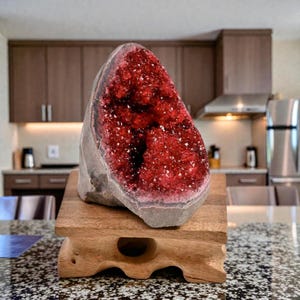 Może przedstawiać: Duża, surowa geoda ametystowa z żywym, czerwonym wnętrzem, wystawiona na małym, rzeźbionym drewnianym stojaku. Zewnętrzna strona geody jest stonowana szara, kontrastująca z błyszczącym kryształowym wnętrzem. Drewniany stojak ma naturalne wykończenie.