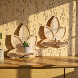 Peut inclure: Deux étagères en bois en forme de fleur de lotus fixées à un mur texturé. Chaque étagère possède une petite plateforme rectangulaire. L'une contient une plante succulente en pot, l'autre un petit récipient en verre avec des plantes. Les étagères sont en bois clair.