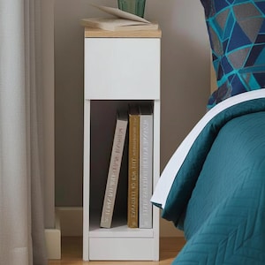 Może przedstawiać: Biały stolik nocny z jasnym drewnianym blatem i otwartą półką. Na półce widoczne są dwie książki. Na górze stos książek i zielona szklanka. Na tle turkusowa narzuta i wzorzysta poduszka.