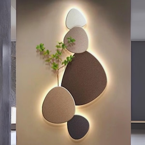 Può includere: Un'opera d'arte murale moderna con una serie di pannelli ovali strutturati a strati in tonalità di marrone, beige e bianco. Una piccola pianta verde è posta al centro. I pannelli sono illuminati da una luce calda e soffusa.