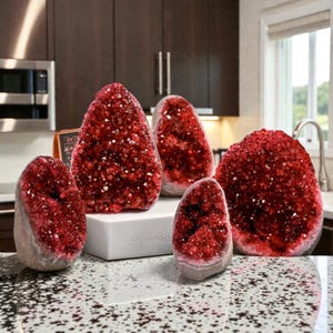 Könnte beinhalten: Fünf rote Kristallgeoden unterschiedlicher Größe werden auf einer weißen Arbeitsplatte ausgestellt. Die Geoden haben eine raue, strukturierte Außenseite und ein funkelndes, kristallines Inneres. Der Hintergrund umfasst eine Küche mit Mikrowelle und Schränken.