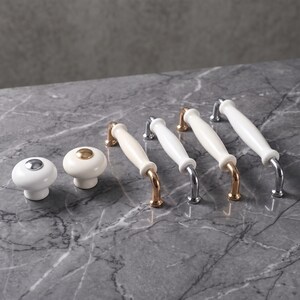 May include: Two round white ceramic knobs with metallic centers and four elongated white ceramic drawer pulls with metallic ends. The hardware features a mix of silver-tone and gold-tone metal finishes, arranged in a row on a gray surface.