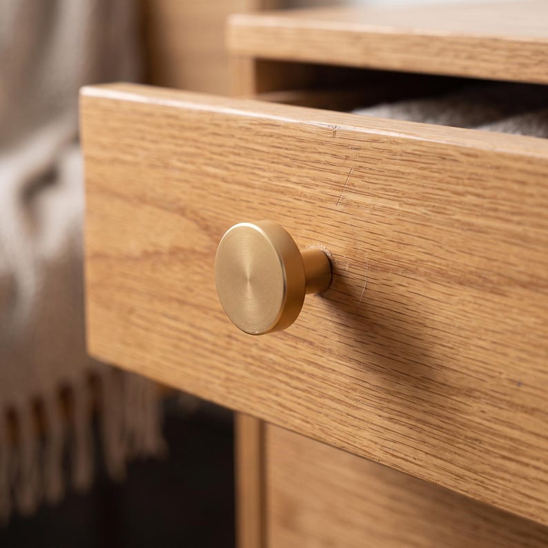 Pu&ograve; includere: Un pomello per mobili circolare in metallo dorato spazzolato fissato su un frontale di cassetto in legno di rovere chiaro. Il pomello presenta una superficie piatta e liscia con una lucentezza metallica, in contrasto con la venatura del legno.