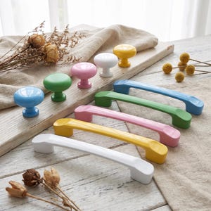 May include: A collection of colorful cabinet hardware including five round knobs and four arched drawer pulls in solid glossy finishes of white, yellow, pink, green, and blue, arranged in rows on a wooden surface.