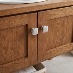 May include: A wooden vanity cabinet featuring a medium brown oak grain finish with two shaker-style doors. Each door is fitted with a square, brushed silver metal knob. A white ceramic sink basin is visible along the top edge of the cabinet.