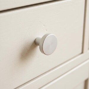 Pu&ograve; includere: Un pomello per mobili circolare in metallo argentato spazzolato fissato su un frontale di cassetto in legno verniciato color bianco sporco. Il pomello presenta una superficie piatta e liscia con una base cilindrica, centrato sul pannello del cassetto.