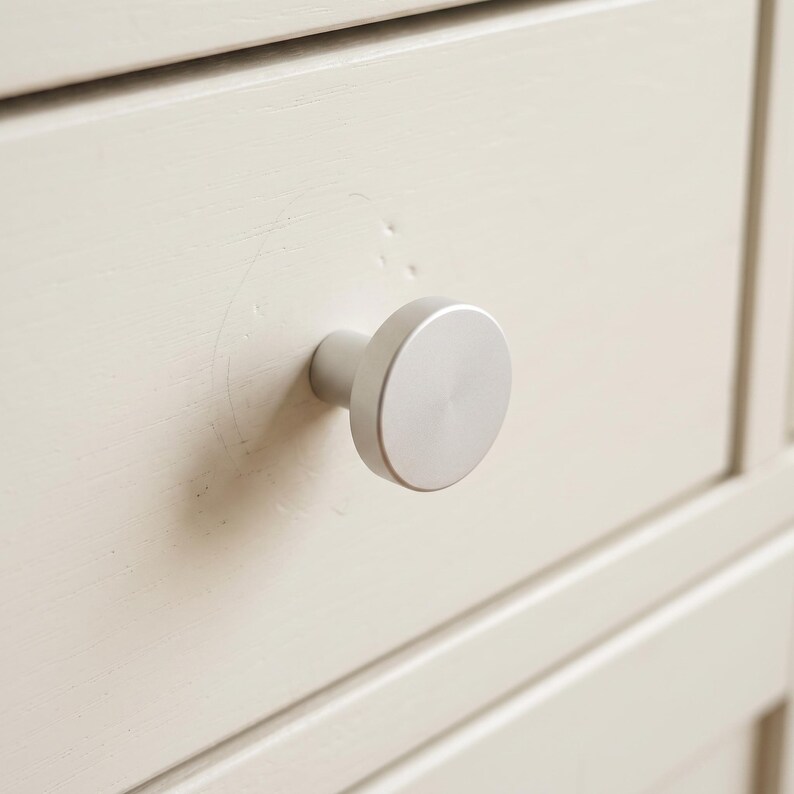 Pu&ograve; includere: Un pomello per mobili circolare in metallo argentato spazzolato fissato su un frontale di cassetto in legno verniciato color bianco sporco. Il pomello presenta una superficie piatta e liscia con una base cilindrica, centrato sul pannello del cassetto.
