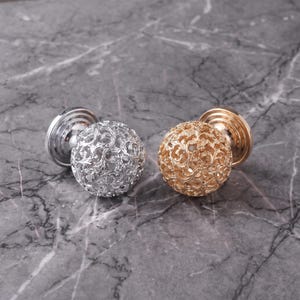 May include: Two spherical cabinet knobs, one in a polished silver finish and one in a gold finish. Each knob features an intricate, openwork filigree pattern of swirling vines and leaves, attached to a circular base plate for mounting.