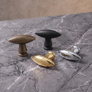 May include: Four cabinet knobs with an elongated, oval-shaped handle and a cylindrical base. The set includes one textured bronze knob, one matte black knob, one polished gold-tone knob, and one polished silver-tone chrome knob.