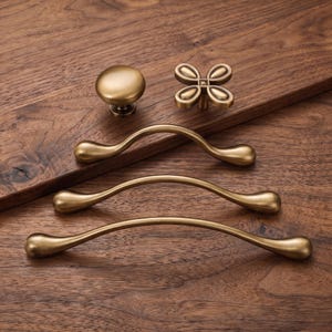May include: A collection of five brass-toned metal hardware pieces including one round knob, one four-petaled flower-shaped knob, and three curved drawer pulls of varying lengths, all featuring a smooth, brushed metallic finish.