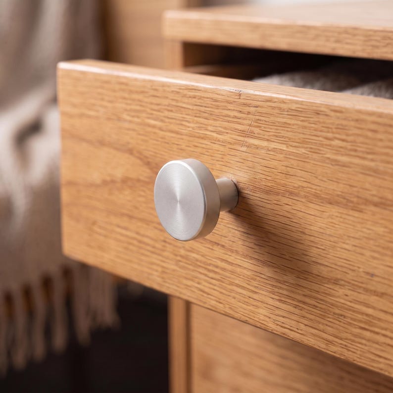 Pu&ograve; includere: Un pomello per mobili circolare in metallo argento spazzolato fissato su un frontale di cassetto in legno chiaro. Il pomello presenta una faccia piatta e liscia con finitura metallica, montato centralmente sulla venatura orizzontale del legno.