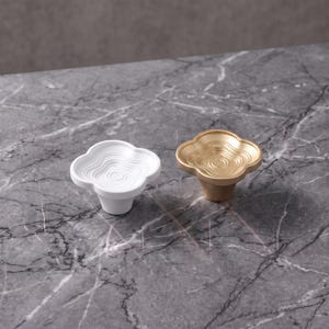May include: Two cabinet knobs, one in matte white and one in metallic gold, each featuring a four-lobed clover shape with a concentric, rippled, topographical pattern on the top surface and a tapered cylindrical base for mounting.