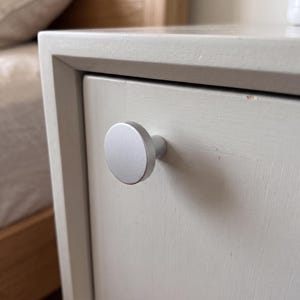 Pu&ograve; includere: Un pomello per mobile circolare grigio chiaro con una finitura opaca liscia, fissato su un frontale di cassetto grigio chiaro. Il pomello presenta una faccia rotonda, piatta e semplice e una base cilindrica, come elemento di ferramenta.