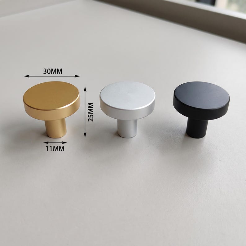 Pu&ograve; includere: Tre pomelli rotondi per mobili con finiture oro, argento e nero. Il pomello dorato presenta etichette con dimensioni che indicano un diametro di 30 mm, un'altezza di 25 mm e una base di 11 mm. Ogni pomello ha una parte superiore piatta e una base cilindrica.