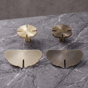 May include: Two circular cabinet knobs and two semi-circular cabinet pulls featuring a sunburst pattern with fine radiating lines. The hardware is finished in two distinct metallic tones, with one set in a bright gold and the other in a dark bronze.
