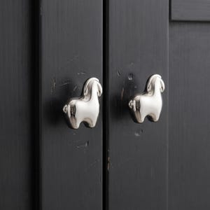 Può includere: Due pomelli per armadietto in metallo argentato lucido a forma di lama o alpaca stilizzati. Ogni pomello presenta una superficie liscia e riflettente ed è montato su un'anta di armadietto in legno grigio antracite con venature verticali.