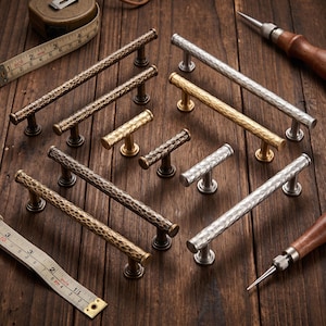 May include: A collection of hammered metal cabinet pulls and T-bar knobs in bronze, brass, and silver finishes. The hardware features a textured surface and cylindrical bars. A measuring tape shows increments in inches for scale reference.