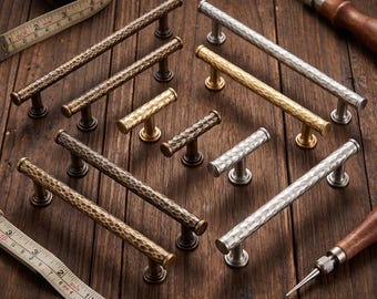 May include: A collection of hammered metal cabinet pulls and T-bar knobs in bronze, brass, and silver finishes. The hardware features a textured surface and cylindrical bars. A measuring tape shows increments in inches for scale reference.