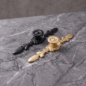 May include: Two decorative cabinet pulls, one in matte black and one in brushed gold. Each pull features a central circular knob with a ridged edge, flanked by elongated, leaf-shaped metal extensions with a smooth, symmetrical design.