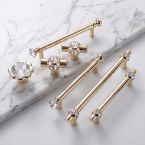 May include: A collection of gold-toned metal cabinet hardware including several bar pulls and knobs. The hardware features clear, faceted crystal accents at the ends and centers. One knob has a flower-shaped base with a large central crystal.