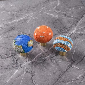 Peut inclure: Trois boutons de placard sphériques aux surfaces texturées. Un bouton est bleu avec des détails blancs et verts, un est orange uni avec des indentations blanches, et un présente des bandes horizontales bleues et orange en relief.