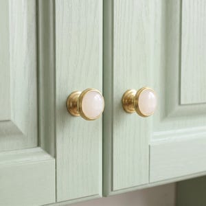 Op de afbeelding: Twee ronde kastknoppen met een gladde, ondoorzichtige witte cirkelvormige voorkant in een basis van geborsteld messing. De knoppen zijn gemonteerd op twee aangrenzende licht saliegroene houten kastdeuren met een zichtbare verticale houtnerf.