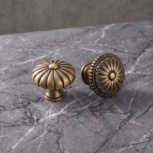 May include: Two round cabinet knobs with an antique brass finish. One knob features a fluted, pumpkin-like design with vertical ridges, while the other displays a detailed, engraved floral pattern with a beaded border around the edge.
