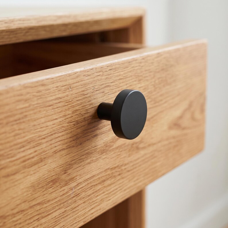 Pu&ograve; includere: Un pomello per mobile circolare nero opaco fissato su un frontale di cassetto in legno di quercia marrone chiaro. Il pomello presenta una faccia rotonda e piatta con una base cilindrica, in contrasto con la venatura del legno naturale.