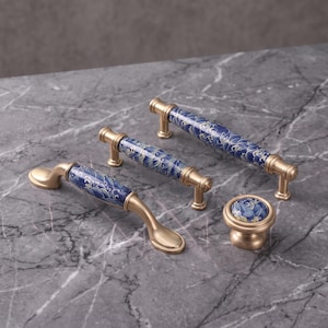 Pode incluir: Um conjunto de ferragens para móveis incluindo dois puxadores longos, um puxador arqueado e um botão redondo. Cada peça apresenta um acabamento em metal dourado escovado com uma inserção central de cerâmica azul com padrão de ondas.