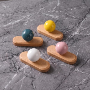 Può includere: Quattro ganci da parete, ciascuno composto da una base in legno liscia e arrotondata con un pomello sferico. I pomelli presentano una finitura lucida in colori solidi: blu ottanio, giallo brillante, rosa tenue e bianco, su legno chiaro.