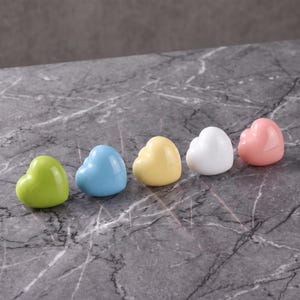 Pode incluir: Cinco objetos lisos em forma de coração dispostos em fila. As cores da esquerda para a direita são verde-limão, azul-claro, amarelo-pálido, branco e rosa-suave. Cada coração tem um acabamento brilhante e reflexivo e uma forma arredondada.