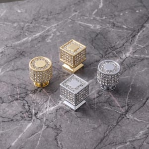 May include: Four cabinet knobs featuring two round and two square shapes. Each knob is finished in either gold or silver tone metal and is encrusted with numerous small, clear, reflective crystals covering the sides and top surfaces.