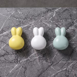 May include: Three smooth, rounded ceramic objects shaped like rabbit heads, arranged in a horizontal row. The left object is yellow, the centre object is white, and the right object is a soft, pale green. Each piece features two small ears.