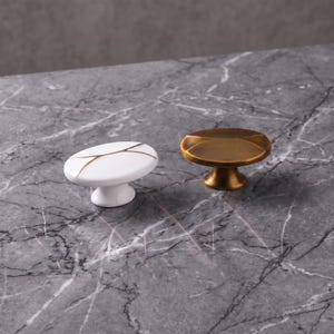 Peut inclure: Deux boutons de placard de forme ovale. Un bouton est blanc avec de fines lignes dorées sur la surface supérieure. Le second bouton est en métal couleur laiton brossé avec un dessus arrondi lisse et une base en piédestal pour la fixation.