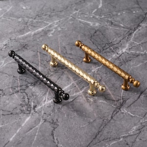 May include: Three cabinet pulls with a textured, diamond-patterned cylindrical bar and spherical ends. One pull is finished in matte black, one in polished gold, and one in a brushed brass tone, each supported by two small cylindrical mounting posts.