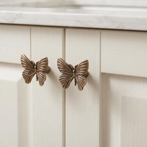 Op de afbeelding: Twee messingkleurige metalen kastknoppen in de vorm van vlinders met gedetailleerde vleugeltexturen. Elke knop is gemonteerd op een lichtgekleurde kastdeur met verticale panelen en dient als functionele handgreep voor het openen en sluiten.